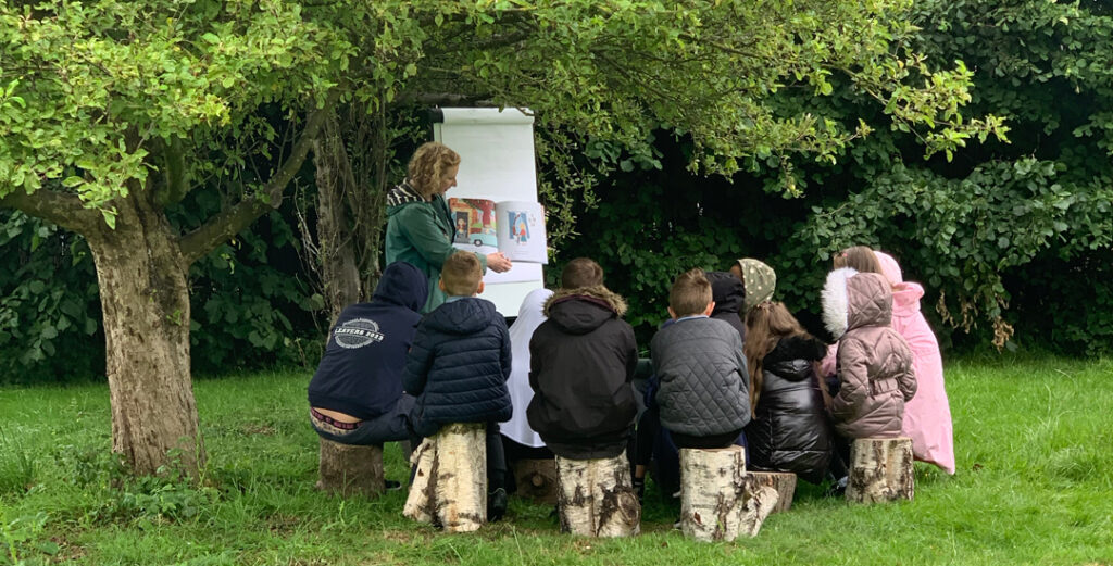 School pupils sat outside under a leafy tree, with KPT patron Lydia Monks doing an illustration session